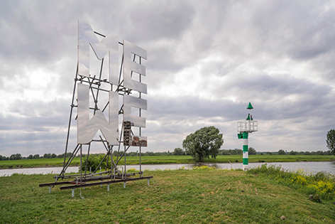 Verspiegelte Lettern ME und WE auf einer Wiese vor einem Fluss bei wolkigem Himmel